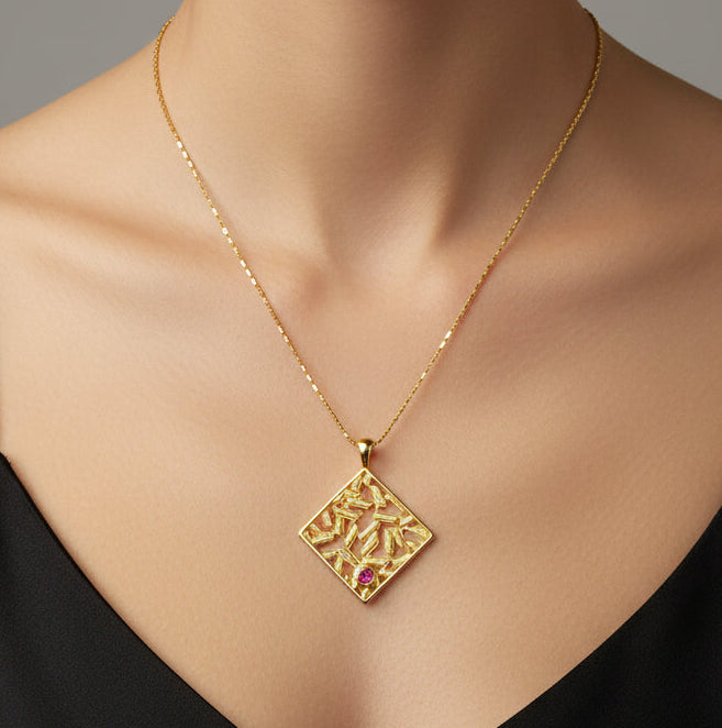 Gold necklace with a square pendant featuring a red gemstone on a light green cushion.
