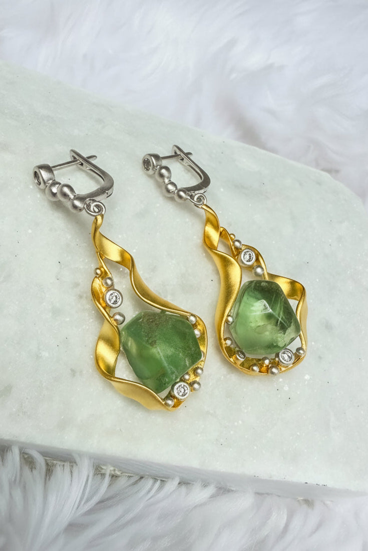 Gold and silver earrings with green gemstones on a white stone surface
